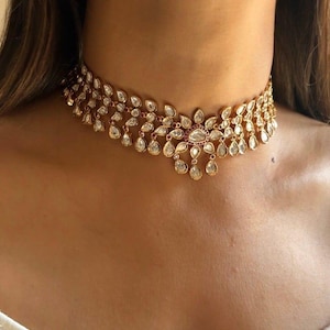 Può includere: Una collana girocollo dorata con un design floreale. La collana presenta molteplici pietre preziose chiare a forma di goccia e un gruppo centrale di pietre più grandi. La collana è indossata intorno al collo.