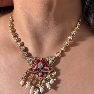 May include: Gold-toned necklace featuring a central pendant with a red enamel design and clear gemstone accents. The pendant is adorned with dangling white pearls. The necklace has a delicate chain with clear gemstone details.