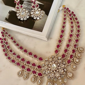 May include: A gold-coloured jewellery set featuring a three-strand necklace and matching earrings. The necklace and earrings are adorned with ruby-coloured stones and clear, sparkling accents. The necklace has a large, floral-shaped pendant.