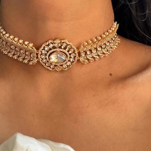 May include: A gold choker necklace featuring a large, clear gemstone at its centre. The necklace is adorned with smaller gemstones and pearls.
