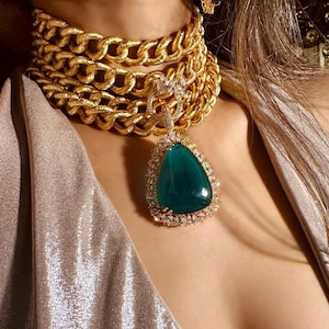 Puede incluir: Un collar de cadena dorada con un gran colgante en forma de lágrima con una piedra de color verde azulado rodeada de pequeñas piedras transparentes. También se ven pendientes a juego. La joya se lleva sobre una tela brillante plateada-dorada.