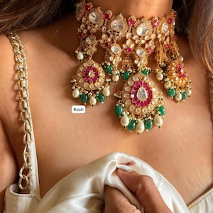 May include: A gold choker necklace featuring pink and white stones, green beads, and a pearl drop. The necklace has a floral design and is adorned with intricate details. The text "Rosh" is visible in the image.