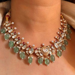 May include: A gold necklace featuring a large, round, clear stone at its centre. The necklace is embellished with smaller, clear stones and red stones. Green beads also adorn the necklace.