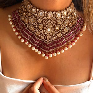 May include: A luxurious gold-toned choker necklace with intricate detailing. The necklace features a central design of clear gemstones and is bordered by a row of deep red gemstones. Small white pearls line the bottom edge.