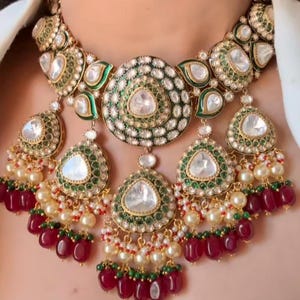 May include: A gold necklace with a large green and white centerpiece, surrounded by smaller green and white pendants. The necklace is adorned with pearls and red gemstones.