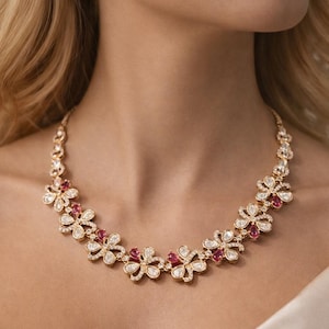 Puede incluir: Un collar y pendientes a juego en tono dorado con un diseño floral. Cada flor tiene piedras preciosas blancas y rojas. La joyería la lleva una persona con cabello rubio y una blusa color crema.