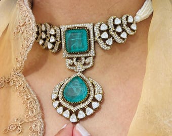 May include: A gold choker necklace with a large emerald green gemstone pendant. The necklace is decorated with smaller white gemstones.