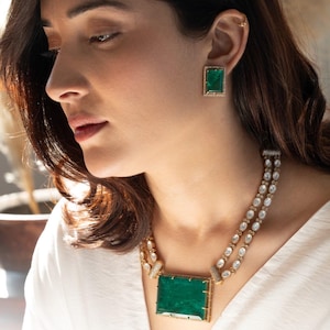 May include: A woman wearing a white top with a gold necklace featuring a large emerald green stone pendant and matching emerald green earrings. The necklace is made of small, clear stones.