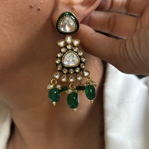 May include: Gold dangle earrings with a large, clear, faceted gemstone surrounded by smaller, clear gemstones. The earrings have a green enamel base and green gemstone drops.