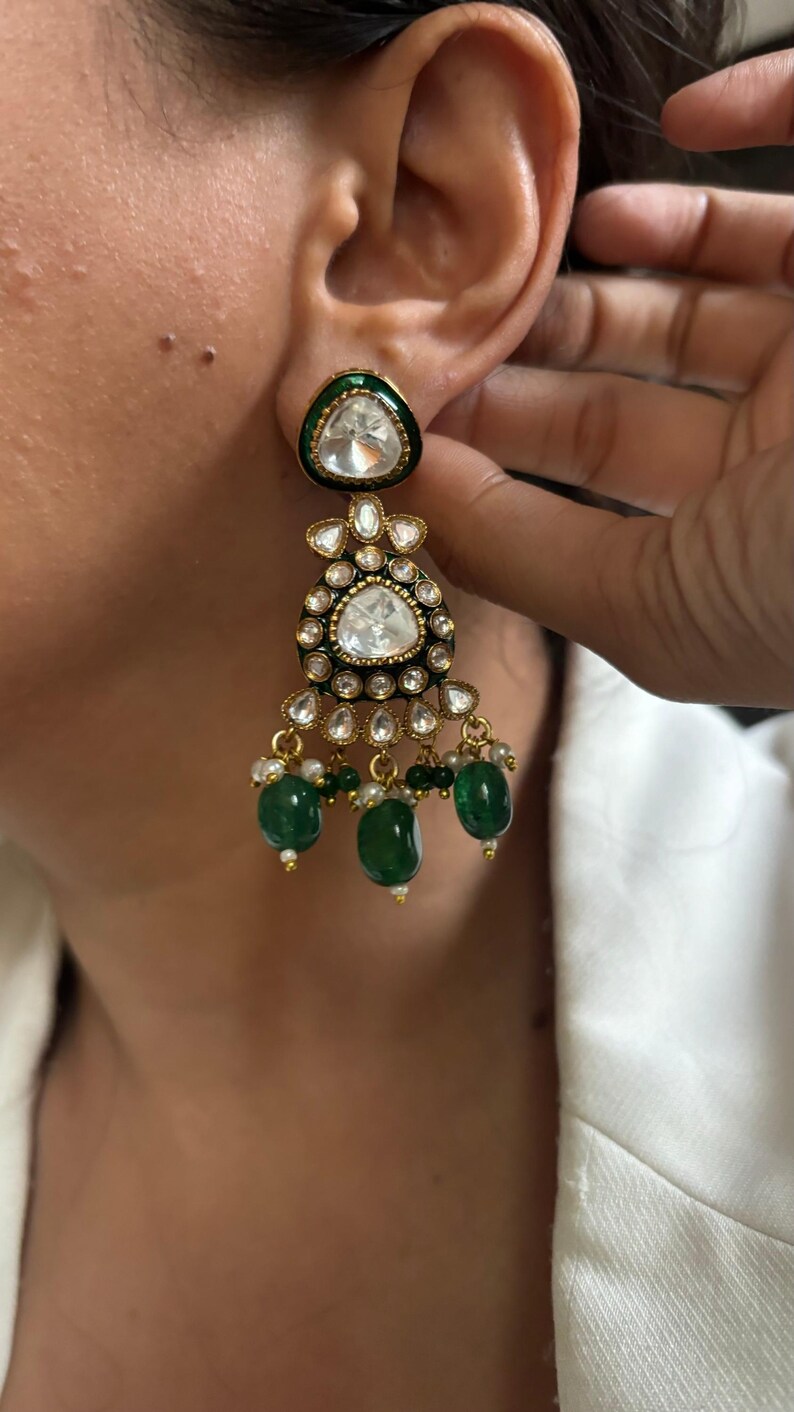 May include: Gold dangle earrings with a large, clear, faceted gemstone surrounded by smaller, clear gemstones. The earrings have a green enamel base and green gemstone drops.