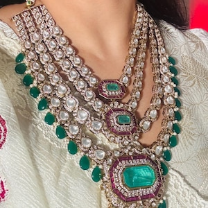 May include: A multi-layered necklace featuring large emerald-cut green gemstones, surrounded by smaller clear stones and red accents. The necklace has multiple strands with teardrop-shaped green gems. The jewelry is set against a white background.