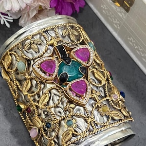 Può includere: Bracciale a polsino in argento con filigrana dorata e pietre preziose colorate. Il bracciale presenta un design centrale con una pietra verde, circondata da pietre triangolari rosa e diamanti. I bordi sono adornati con corda dorata e dettagli di foglie.
