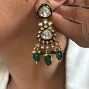 May include: Gold earrings with a large, clear, faceted gemstone surrounded by smaller gemstones. The earrings have a green enamel base and dangle with green and white beads.