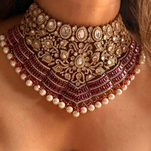 May include: A luxurious gold-toned choker necklace with intricate detailing. The necklace features a central design with clear and pink gemstones, surrounded by a border of red stones and white pearls. A statement piece for special occasions.