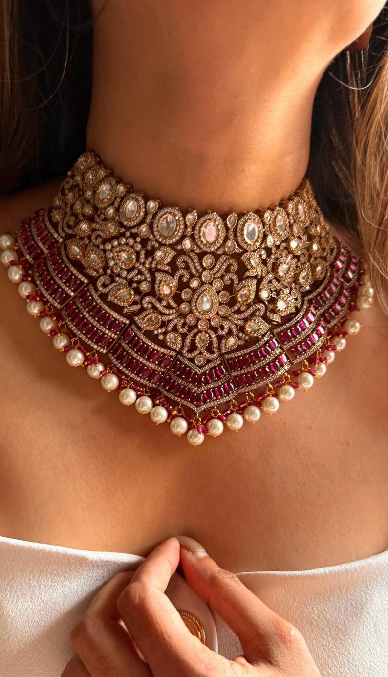 May include: A luxurious gold-toned choker necklace with intricate detailing. The necklace features a central design with clear and pink gemstones, surrounded by a border of red stones and white pearls. A statement piece for special occasions.