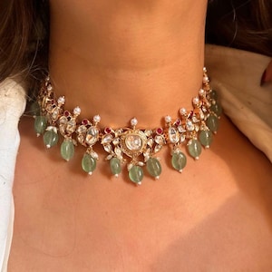 May include: A gold choker necklace with a large, round, clear stone in the center. The necklace is decorated with smaller clear stones, green stones, and red stones.