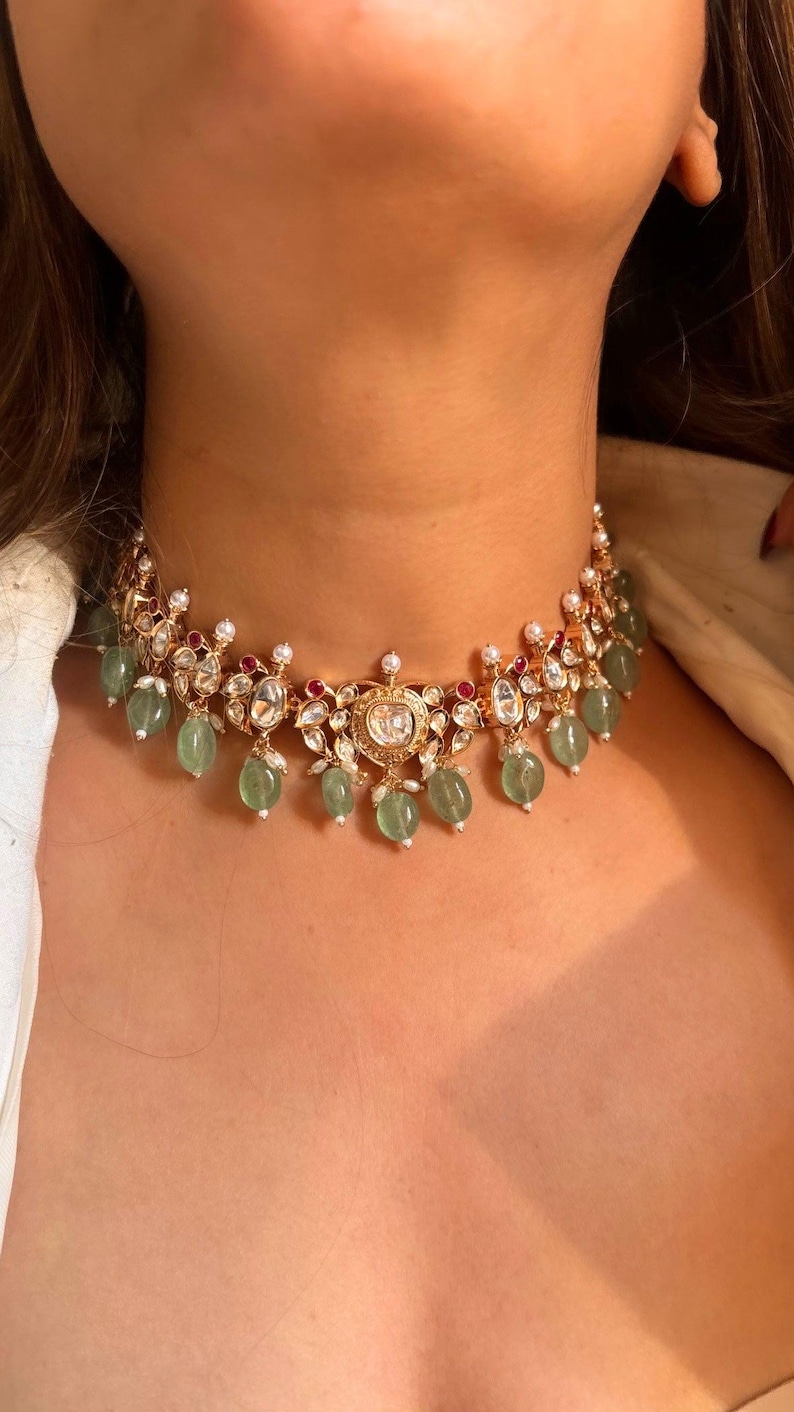 May include: A gold choker necklace with a large, round, clear stone in the center. The necklace is decorated with smaller clear stones, green stones, and red stones.