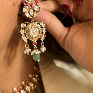 May include: Gold earrings with a large, clear gemstone surrounded by smaller gemstones. The earrings have a teardrop shape and dangle from a floral design. The earrings are accented with small pearls and green gemstones.