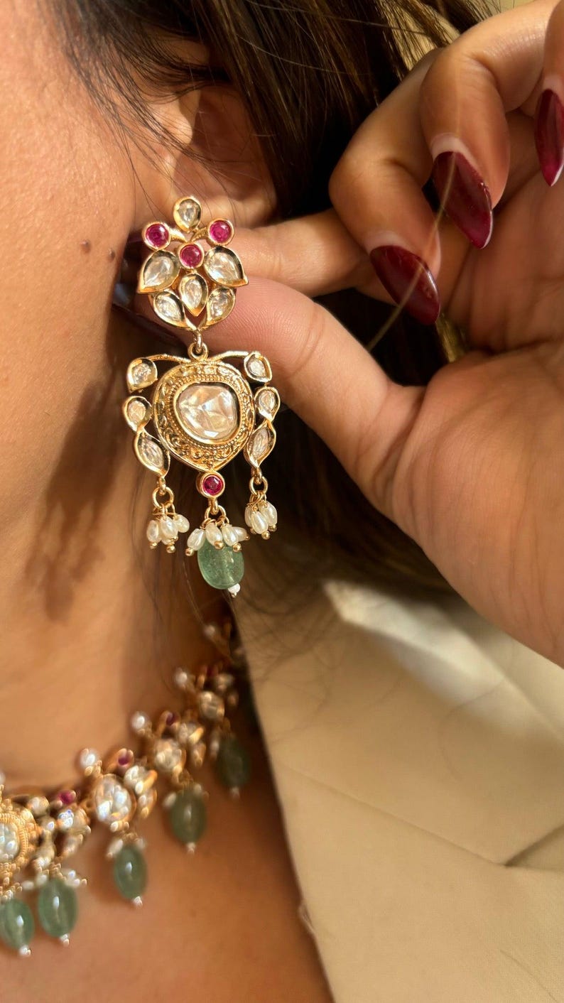 May include: Gold earrings with a large, clear gemstone surrounded by smaller gemstones. The earrings have a teardrop shape and dangle from a floral design. The earrings are accented with small pearls and green gemstones.