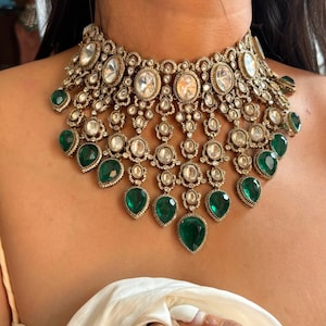 Peut inclure: Un collier en or et argent avec de grandes pierres claires et des pierres vertes en forme de larme. Le collier est superposé et présente un design délicat.
