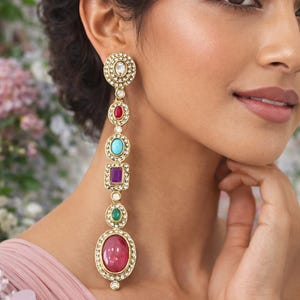 May include: Long, gold-coloured dangle earrings with a sequence of colourful gemstones. The earrings feature a large, round, ruby-coloured stone at the bottom, with other stones in red, turquoise, purple, and green. The earrings are adorned with small, clear crystals.