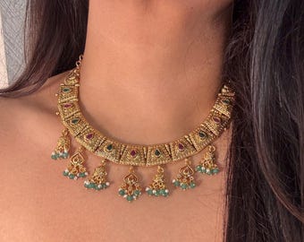 May include: A gold-toned necklace featuring a series of rectangular, textured metal links. Each link is inlaid with a small, teardrop-shaped red or green stone. Dangling from the bottom are several small, bell-shaped charms with green beads.