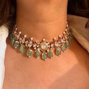 May include: A gold choker necklace with a large, round, clear stone in the center. The necklace is decorated with smaller clear stones, green stones, and red stones. The necklace is on a person's neck.