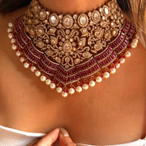 May include: A gold-toned choker necklace with intricate detailing. The necklace features clear and ruby-colored stones, with a pearl-lined edge. The design includes floral and geometric patterns, creating a luxurious and ornate appearance. The necklace is worn around the neck.