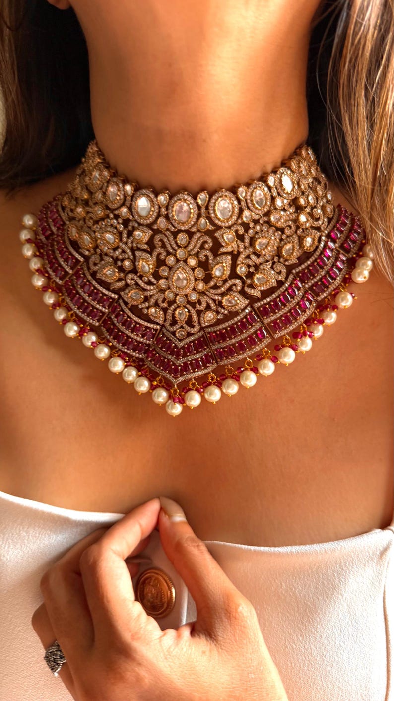 May include: A gold-toned choker necklace with intricate detailing. The necklace features clear and ruby-colored stones, with a pearl-lined edge. The design includes floral and geometric patterns, creating a luxurious and ornate appearance. The necklace is worn around the neck.