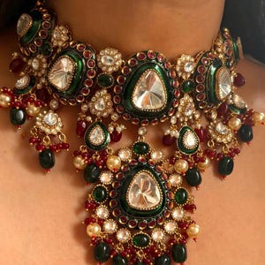 Op de afbeelding: Een gouden ketting met een groen en rood emaille ontwerp. De ketting heeft grote, heldere stenen en kleinere, groene en rode stenen. De ketting heeft een hanger met een grote, heldere steen.