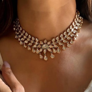 May include: A gold choker necklace with a teardrop-shaped design. The necklace is adorned with clear and green gemstones.