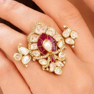 Puede incluir: Un conjunto de anillo y pendientes dorados. El anillo presenta un gran rubí central rodeado de piedras claras en forma de lágrima. Los pendientes tienen un diseño similar. Las joyas están engastadas con pequeñas piedras claras.