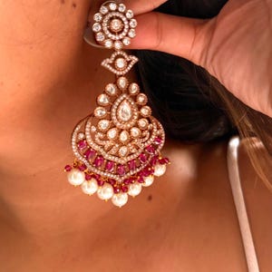 May include: An ornate gold-toned earring with a floral top, a teardrop-shaped center, and a cascading design. The earring features clear and ruby-colored stones, with a fringe of white pearls at the bottom. The earring is being held up by a hand.