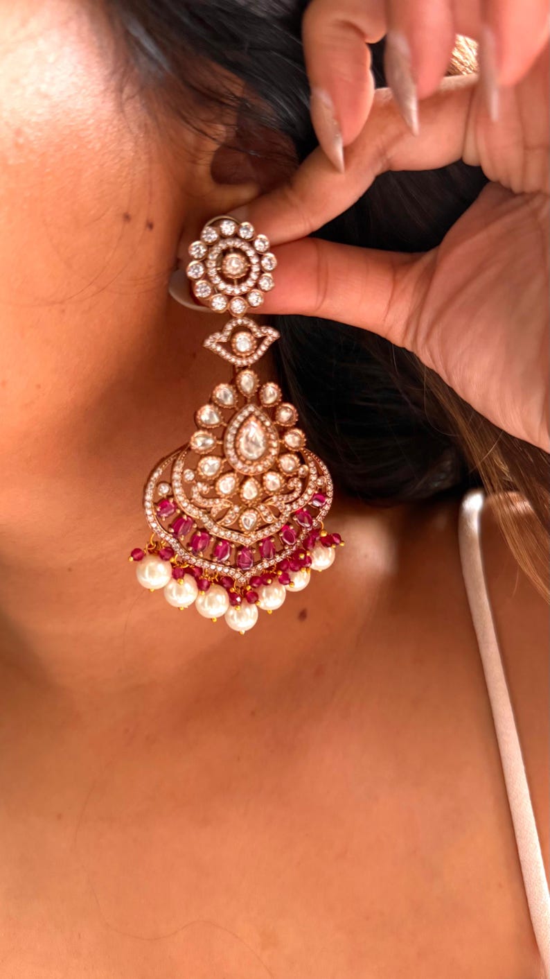 May include: An ornate gold-toned earring with a floral top, a teardrop-shaped center, and a cascading design. The earring features clear and ruby-colored stones, with a fringe of white pearls at the bottom. The earring is being held up by a hand.