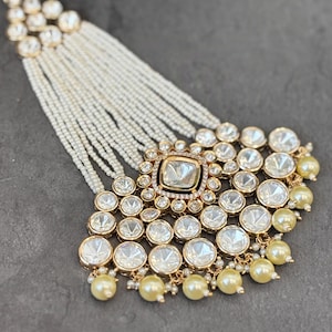 Peut inclure: Coiffe dorée ornée d'une pierre transparente carrée centrale, entourée de petites pierres transparentes. Le design comprend des fausses perles pendantes et des brins de petites perles blanches, créant un look luxueux.