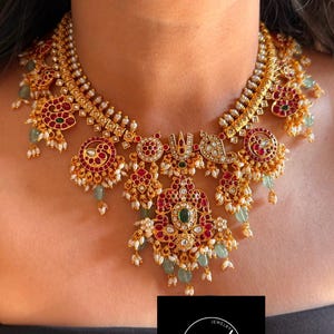 May include: A gold-toned necklace with intricate detailing, featuring red and green gemstones, and small white pearls. The necklace has a central pendant and multiple decorative elements. The jewelry is displayed on a person.