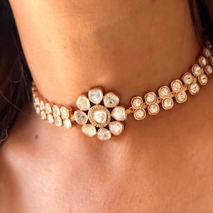 Puede incluir: Un collar gargantilla de oro con un gran colgante de forma de flor transparente. El colgante está hecho de varias piedras más pequeñas engastadas en un marco de oro. El collar está hecho de una cadena de piedras más pequeñas transparentes.