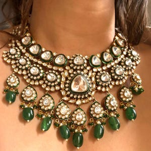 May include: A gold and emerald green statement necklace with a large, ornate design. The necklace features multiple layers of round and teardrop-shaped stones, with a central teardrop-shaped stone.