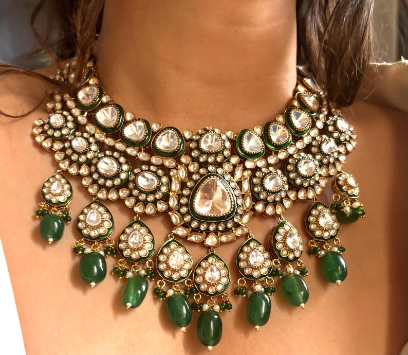 May include: A gold and emerald green statement necklace with a large, ornate design. The necklace features multiple layers of round and teardrop-shaped stones, with a central teardrop-shaped stone.