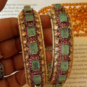 Puede incluir: Dos brazaletes ornamentados con piedras preciosas verdes de talla esmeralda, pequeñas gemas rojas y detalles dorados. Los brazaletes están engastados con cuentas blancas y probablemente están hechos de oro o metal dorado. Las joyas se muestran sobre un fondo borroso.