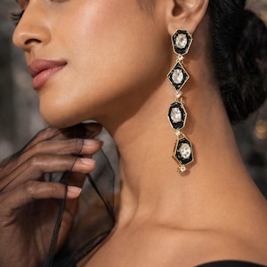 May include: Elegant drop earrings with a geometric design. The earrings feature black enamel and clear, sparkling stones set in a gold-tone metal. The earrings have a long, linear shape with a diamond and hexagon pattern.