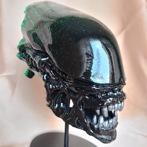 May include: A glossy black Alien head sculpture with green speckles, mounted on a black metal stand. The sculpture features detailed facial features, including sharp teeth and a prominent forehead. The base is round and flat.