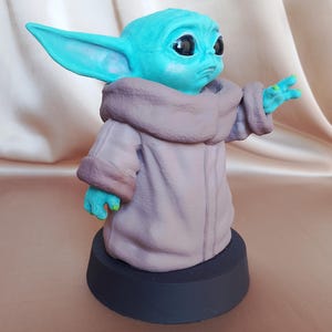 K&ouml;nnte beinhalten: Eine Figur aus teal-gr&uuml;nem und braunem Kunststoff, die einen Charakter aus dem Star Wars-Franchise darstellt. Die Figur tr&auml;gt eine braune Robe und hat gro&szlig;e Ohren. Die Figur steht auf einem schwarzen Sockel.