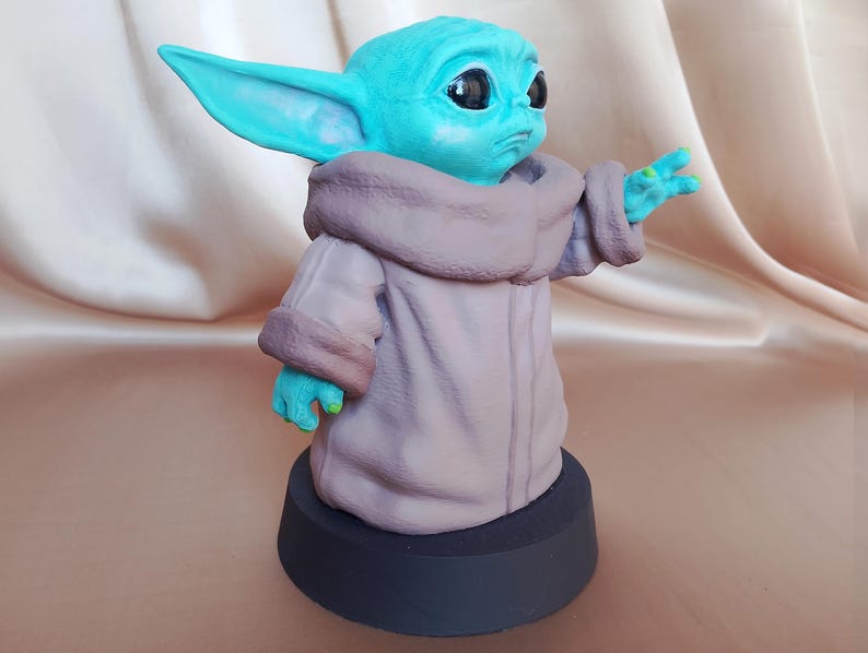 K&ouml;nnte beinhalten: Eine Figur aus teal-gr&uuml;nem und braunem Kunststoff, die einen Charakter aus dem Star Wars-Franchise darstellt. Die Figur tr&auml;gt eine braune Robe und hat gro&szlig;e Ohren. Die Figur steht auf einem schwarzen Sockel.