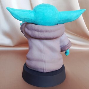 K&ouml;nnte beinhalten: Eine kleine, gr&uuml;ne und braune Figur eines Charakters aus dem Star Wars-Franchise. Die Figur steht auf einem schwarzen Sockel.