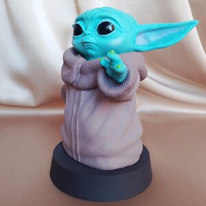 K&ouml;nnte beinhalten: Eine kleine, blaue und gr&uuml;ne Figur eines Charakters aus dem Star Wars-Franchise, die eine braune Robe tr&auml;gt. Die Figur steht auf einem schwarzen Sockel.