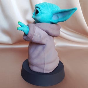 K&ouml;nnte beinhalten: Eine kleine, blaue und gr&uuml;ne Figur eines Charakters aus dem Star Wars-Franchise, die eine braune Robe tr&auml;gt. Die Figur steht auf einem schwarzen Sockel.