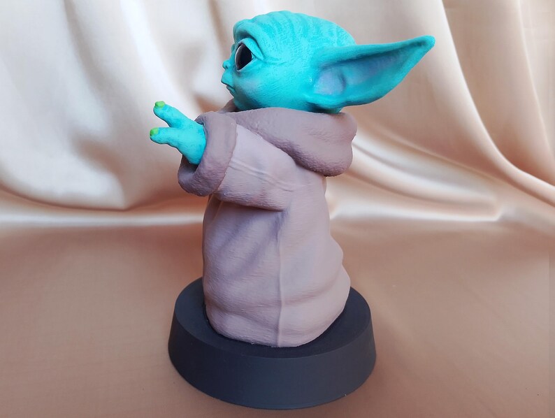 K&ouml;nnte beinhalten: Eine kleine, blaue und gr&uuml;ne Figur eines Charakters aus dem Star Wars-Franchise, die eine braune Robe tr&auml;gt. Die Figur steht auf einem schwarzen Sockel.