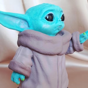 K&ouml;nnte beinhalten: Eine kleine, blaue und gr&uuml;ne Figur eines Charakters aus dem Star Wars-Franchise. Die Figur tr&auml;gt eine braune Robe und hat gro&szlig;e Ohren.