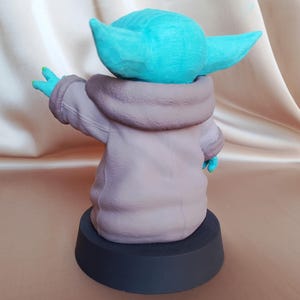 K&ouml;nnte beinhalten: Eine 3D-gedruckte Figur eines Charakters aus dem Star Wars-Franchise, bekannt als "The Child" oder "Baby Yoda". Die Figur ist in hellblau bemalt und tr&auml;gt eine braune Kapuzenrobe. Die Figur steht auf einem schwarzen runden Sockel.